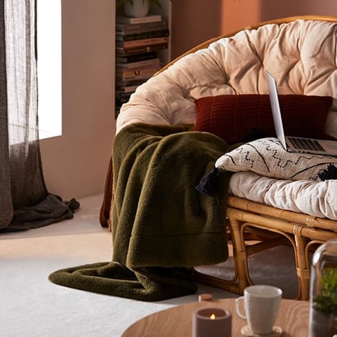 Two-seater rattan chair, with cushions, a laptop on the chair and a dark linen curtain in the background. 
