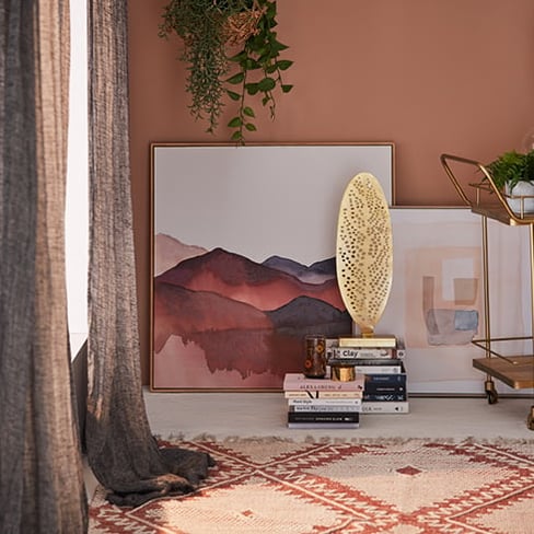 The corner of a room with two warm hued artworks leaning against each other on a wall and 2 stacks of books in-front. 