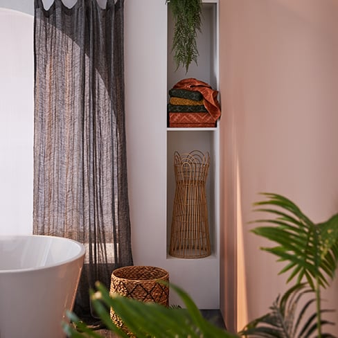 The corner of a bathroom featuring shelving with a plant stand, rattan basket, stack of warm-hued towels.