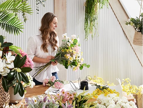 Woman is seen at a table curating her bunch of  colourful flowers and stems.