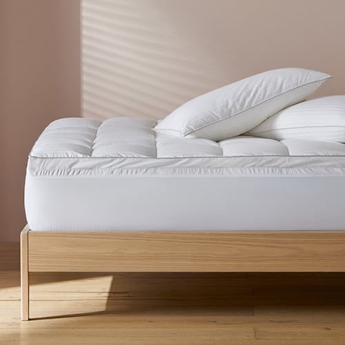 A side-on image of a bed featuring a mattress topper with two pillows stacked on top sitting against a neutral wall.
