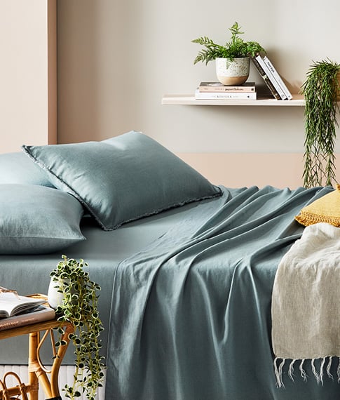 Side-on-view on Bamboo Linen Sage Sheets and matching pillowcases on a bed, next to a rattan side table with books.