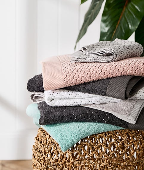 Stack of Bamboo Navara Towels including pink, coal, grey marle, white, and more – all on top of a rattan table.