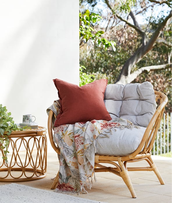 Outdoor scene with bamboo light brown coffee table and chair, dark orange and white cushions and floral throw
