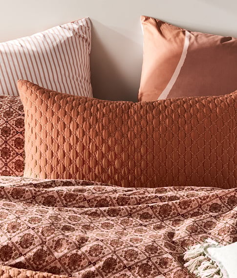 Pillows in a range of terracotta hues, soft and dark pinks leaning on each other on the wall at the top of a bed.  