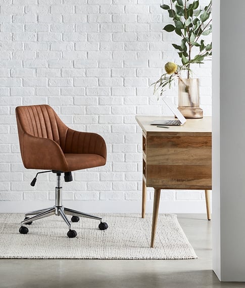 Side-on-view of the Anjuna Natural Desk with a matching desk chair and large rug against a white brick wall. 