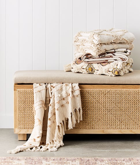 Rattan bench seat styled with throw peeping out from the top of the bench set, plus stack of textured throws on top. 