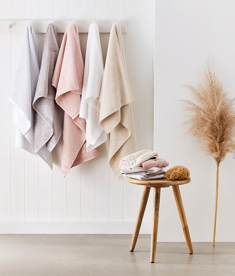Selection of five towels hanging in a row in neutral colours – with a small table in-front with matching facewashes stacked on top.