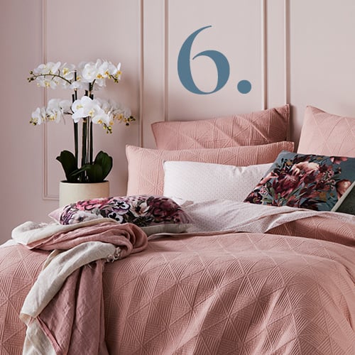 Bedroom with pink bedlinen styled with floral cushions and pink throw, and large orchid on the bedside table. 