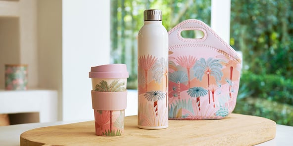Karina Jambrak x Adairs Reusable Mug next to a matching water bottle and lunchbox on a bench.