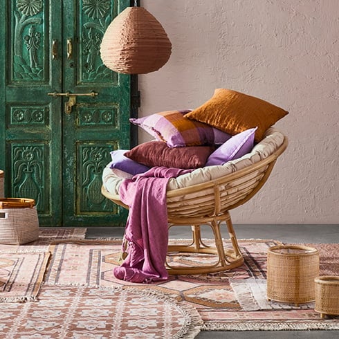 Papasan natural rattan chair styled with warm hued cushions and basket in the background. 