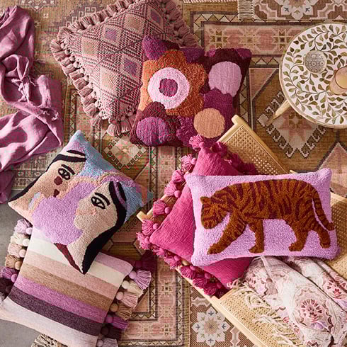 Bird’s eye view of rugs styled on top of each other and warm pink printed, plain, textured and tasselled cushions styled on top. 