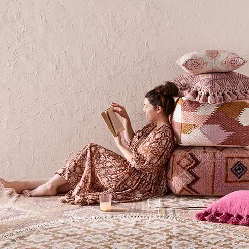 Side-on-view of woman/man resting against a pile of floor and styling cushions in warm hues. 
