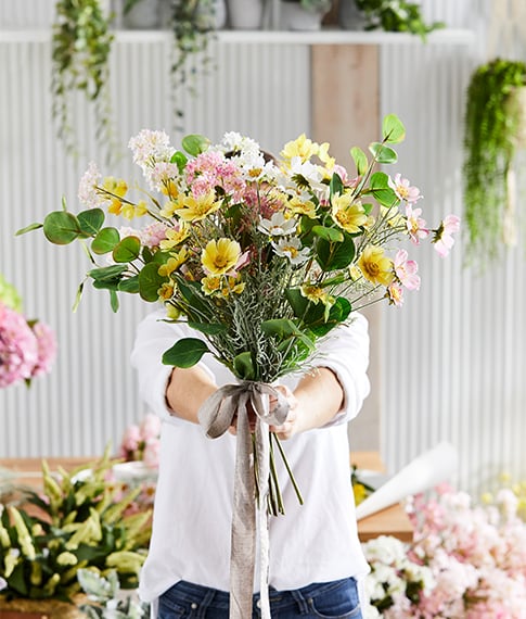 Hands hold a colourful bunch of wildflowers, in shades of light pink and yellow amongst fresh green leaves.