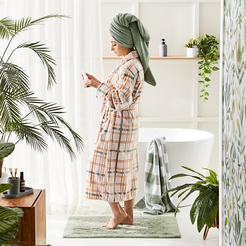 Woman/man in a bathrobe and towel on her head in a bathroom styled with green bathmat, towels, plants, and grey accessories.