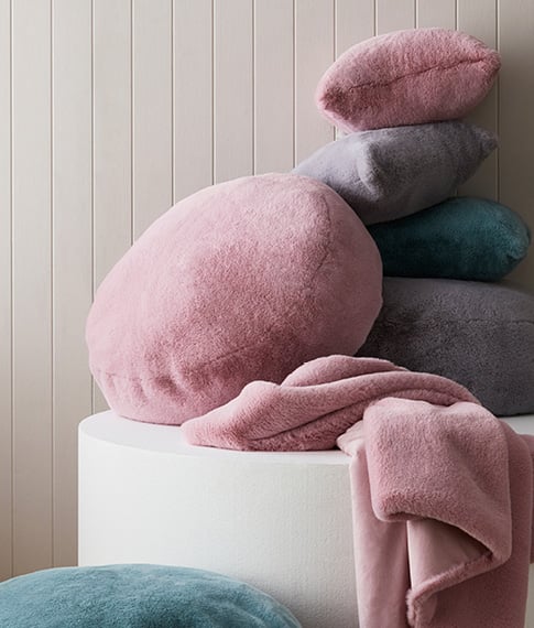 Stack of faux fur floor cushions and styling cushions in purple, blue and pink, as well as a matching pink blanket. 