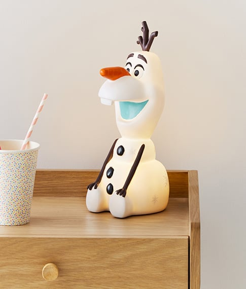Adairs Kids Disney Frozen II Olaf Night Light turned on siting on a natural wooden bedside table next to a spotty printed cup. 