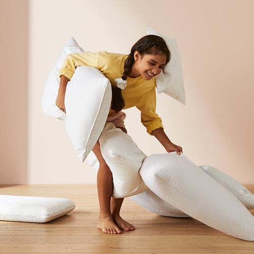 Child smiling standing holding a pillow and surrounded by pillows. 
