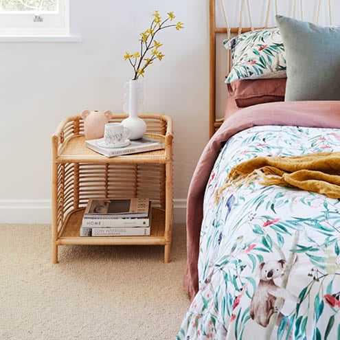 Rattan side table - bottom shelf has books, top features two vases, mug and saucer on top of book next to bed.