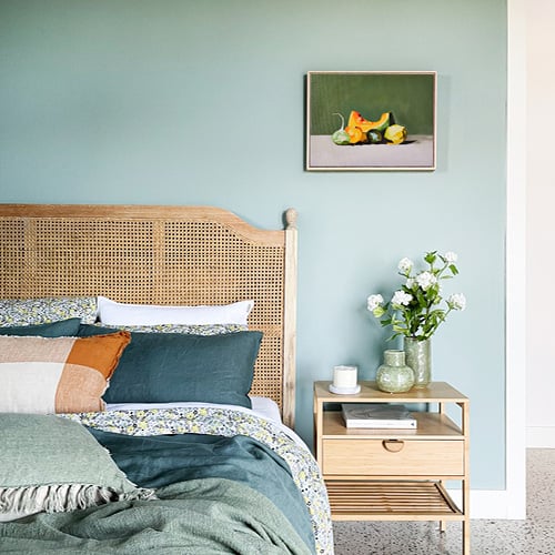 Bed styled with green hued bedlinen, matching cushion and throw on a bed with a rattan bedhead and a matching side table with one drawer.