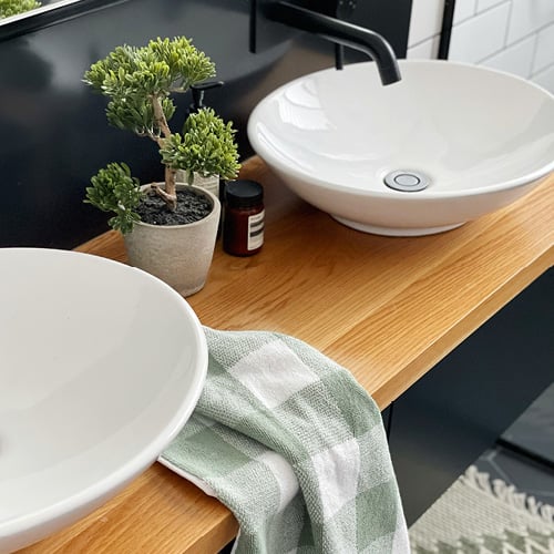Close-up of bathroom sink with white basin, natural wooden top and green towel styled on top next to a small potted plant.