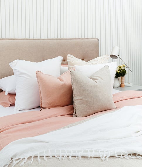Angled view of a bed with neutral and white bedlinen, with matching pillows and styled with Belgian Vintage Cushions in linen and pink.  