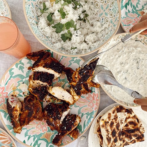 Bird’s eye view of tandoori chicken, rice, raita, and naan bread styled on warm coloured platters and bowls.