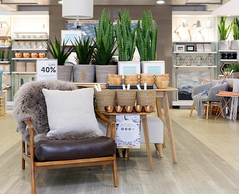 Brown one seater chair with a fury grey throw and a white cushion. The background is a adairs sore setting with various home accessories such as plants and bowls