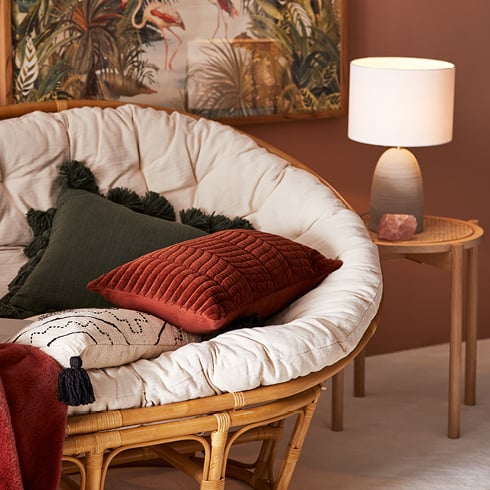 Rattan two-seater chair with tasselled and velvet cushions stacked on the couch next to a side table and lamp.