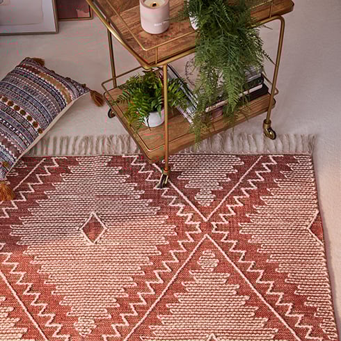 A bar cart featuring books, a candle, and plants, situated on the edge of a red rug with white detailing and tassels. 