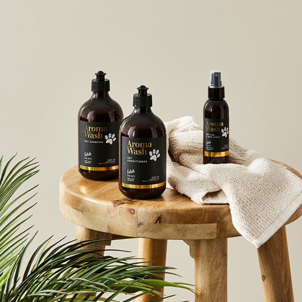 Three Aroma Wash products are arranged on top of a wooden stool with a towel. A large plant sits at the bottom left corner of the frame.