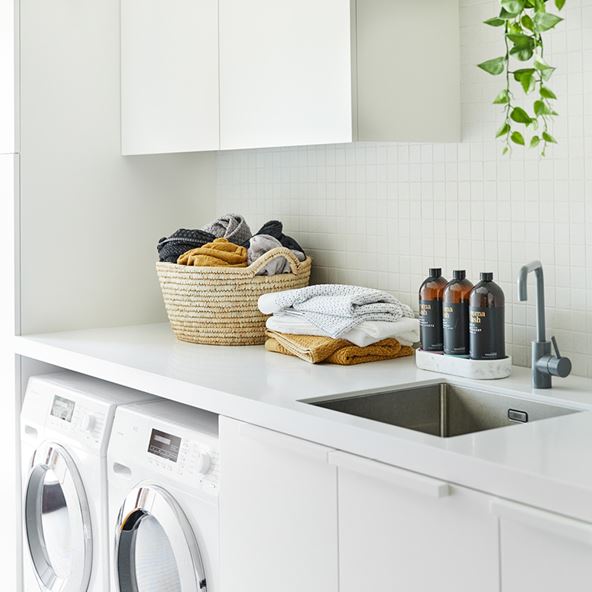 A simple laundry scene with white benchtops and a tiled wall, with a basket of laundry and a pile of towels. Towards the right of the frame, next to a sink, is a marble tray housing three bottles of Aroma Wash laundry liquid.