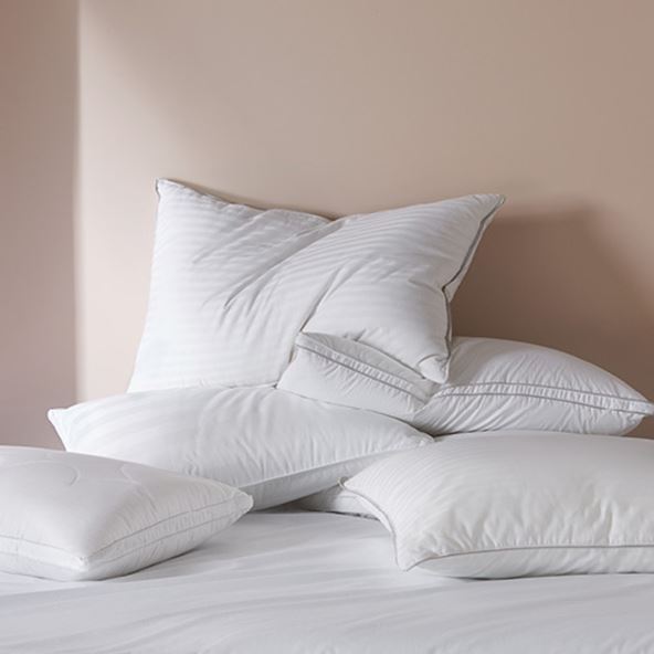 A bed with a selection of various sized pillows stacked on top of each other against a neutral wall. 