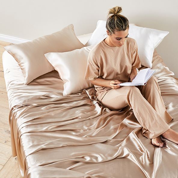 Woman/man sitting up in bed reading on top of pure silk sheets and matching pillowcases in champagne and white.