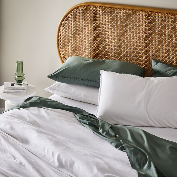 Angled view of a bed with rattan bedhead, side table, and the bed is styled with bamboo bedlinen in white and green.  