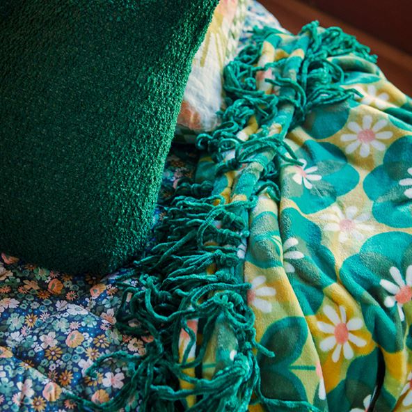 A close up image of the corner of a bed, with a textured green cushion, quilted floral sleeping bag and floral blanket with tasselled edges.