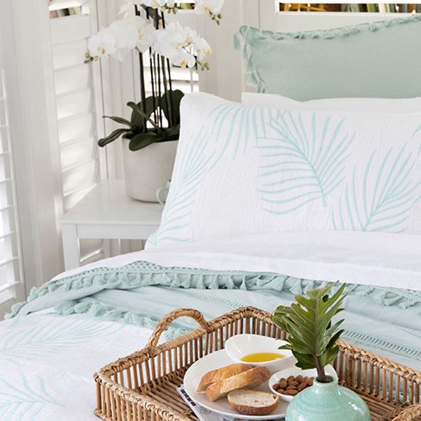 A rattan tray sits on top of a coastal inspired bed and in the background a beautiful white orchid frames the space. 