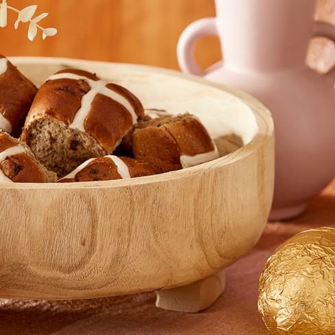 A close up shot of a wooden bowl filled with hot cross buns on a gingham table cloth. Golden easter eggs are scattered around the bowl and in the background is a white vase.