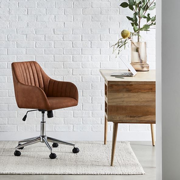 Side-on-view of the Anjuna Natural Desk with a matching desk chair and large rug against a white brick wall. 