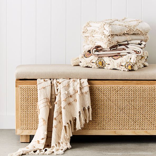 Rattan bench seat styled with throw peeping out from the top of the bench set, plus stack of textured throws on top. 