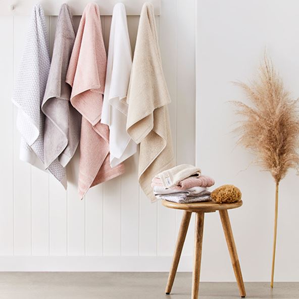 Selection of five towels hanging in a row in neutral colours – with a small table in-front with matching facewashes stacked on top.