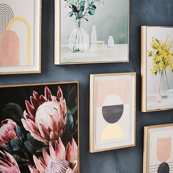 Close-up of a selection of wall art featuring pink, yellow, blue, gold and florals placed at different heights hanging on a dark blue wall. 