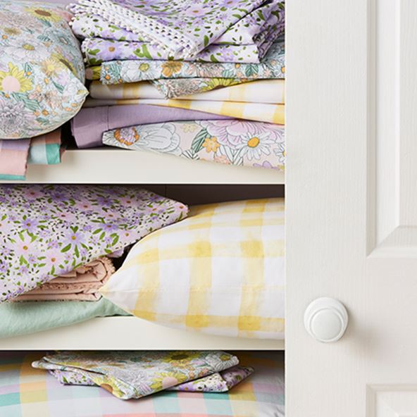 White linen cupboard partly open featuring bedlinen and pillows in pastels including plain, checks and floral patterns. 