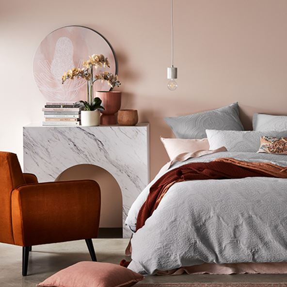 Front-on-view of bedroom with neutral bedlinen, coloured cushions, throws, orange velvet armchair and accessories in warm hues. 