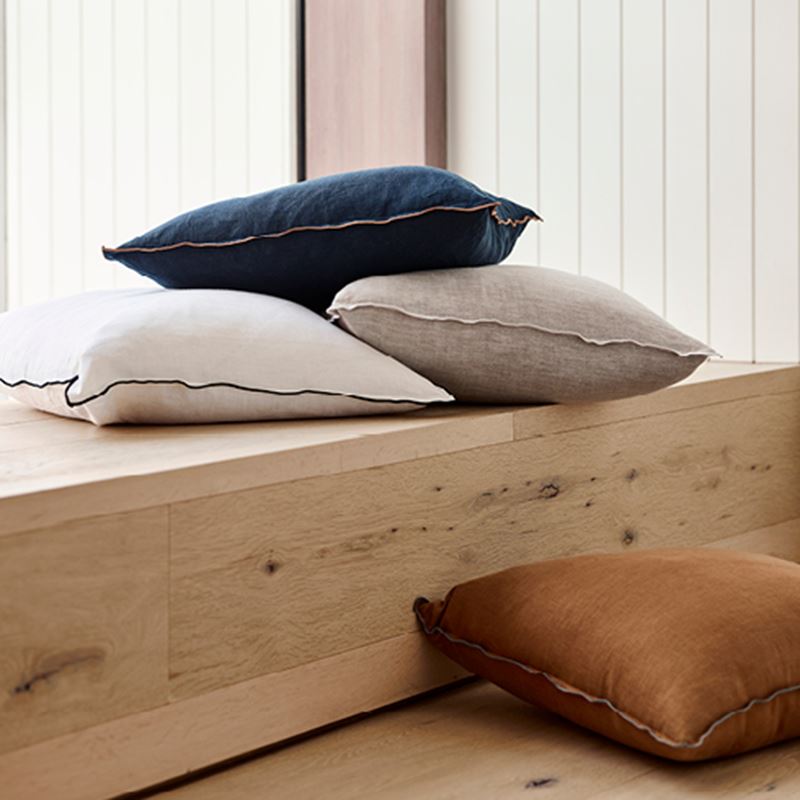 Collection of Mark Tuckey cushions sacked on top of each other sitting on top of a wooden bench seat, with one at the foot of the seat. 