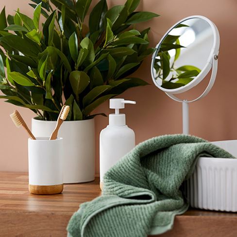 Bathroom scene of a green hand towel, small mirror, soap dispenser, and bamboo tumbler with toothbrushes sit next to a sink.