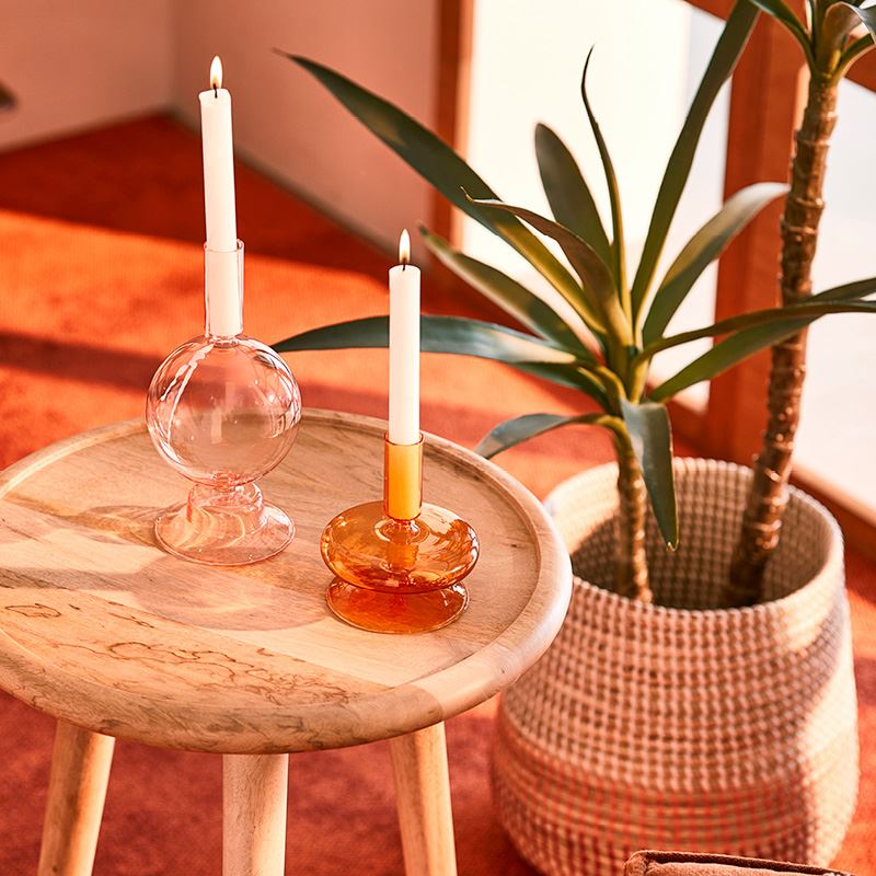 A wooden table with glass candle holders, with a plant in a woven basket to he right. The background is fuzzy orange carpet and the corner of a room.