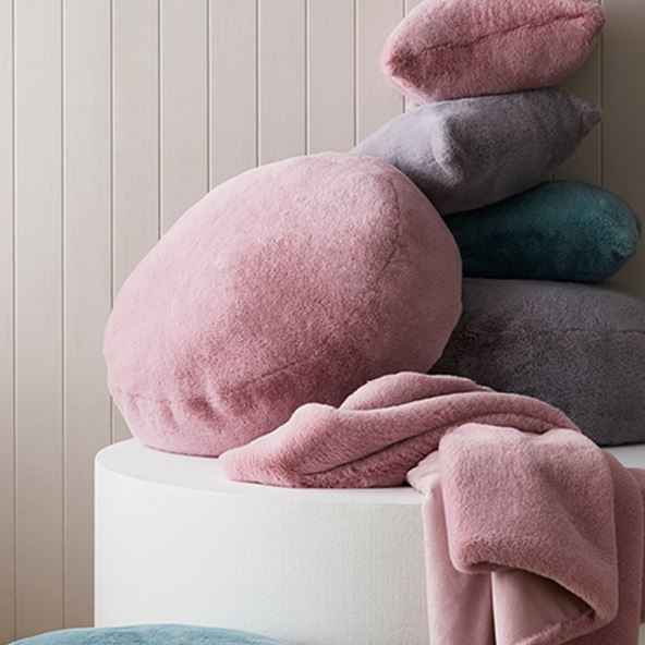 Stack of faux fur floor cushions and styling cushions in purple, blue and pink, as well as a matching pink blanket. 