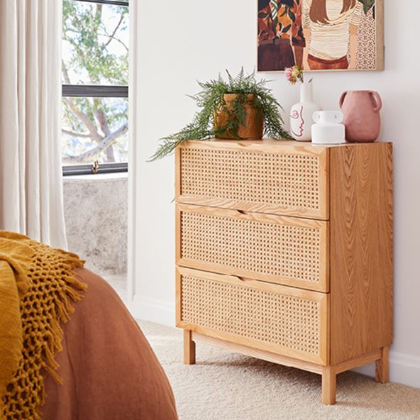 At the end of a bed is an Arden Natural Rattan Chest of 3 Drawers, with vases and a pot plant on top, wall art hanging above. 