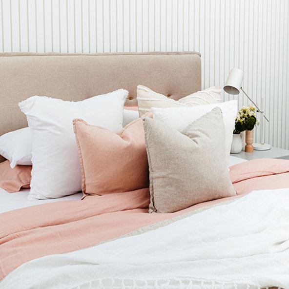 Angled view of a bed with neutral and white bedlinen, with matching pillows and styled with Belgian Vintage Cushions in linen and pink.  